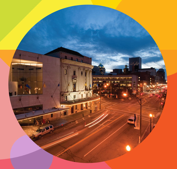 A city street scene at dusk shows a prominently lit classical-style building on the left, possibly a theater or museum. The streets are busy with light trails from moving vehicles. Surrounding buildings are visible under a vibrant twilight sky. The image is framed with rainbow colors.