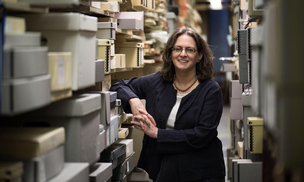 Melissa Mead smiling and leaning against shelf of gray archival boxes in archive.