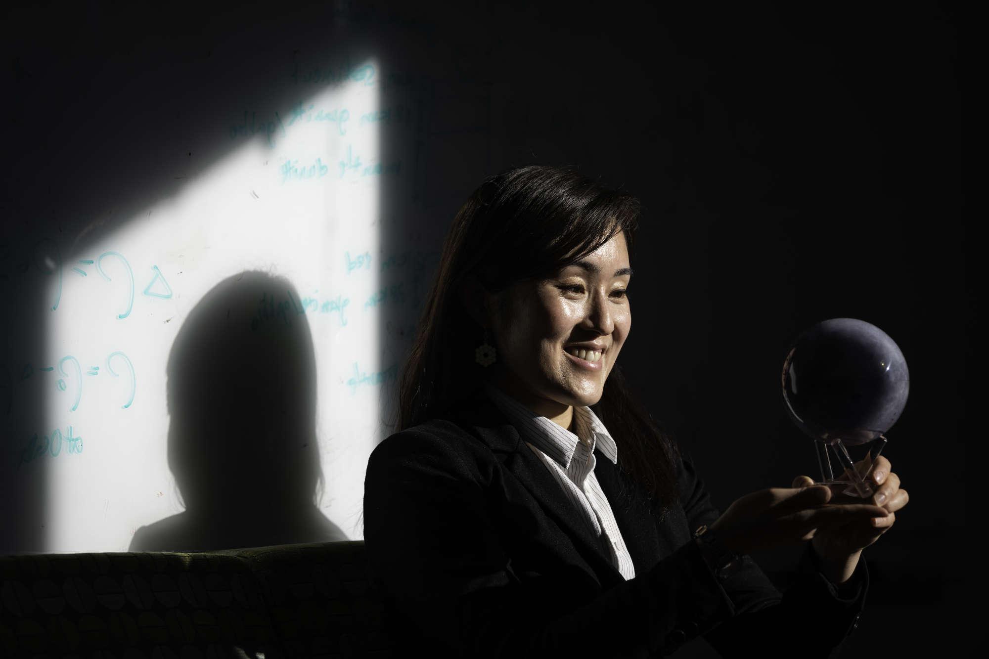 Niki Makajima smiles and looks at a replica moon sculpture while her shadow falls on a whiteboard with equations on it behind her.